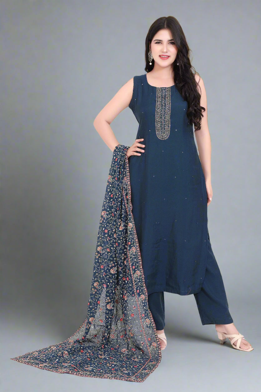 Woman wearing a navy blue kurta with a floral dupatta, standing next to a vase against a beige wall.